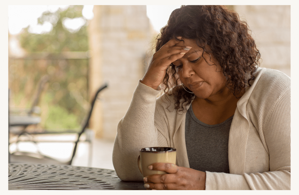 photo of woman sitting at table with coffee mug. She has her hand on her head and looks to be in deep worried thoughts. Canva download with border.